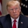 Donald Trump sits at a formal meeting table in a dark suit and red tie, with US flags blurred behind him.